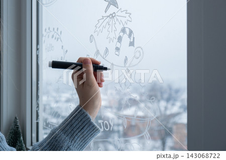 Creative enthusiastic unrecognizable woman in sweater paint window with white marker. Christmas tree decoration on big clear window. Snowy winter outside 143068722