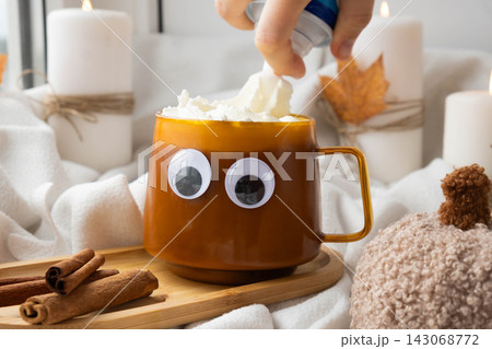Adding whipped cream Funny halloween decoration face with eyes on cup of Pumpkin spiced latte morning warming coffee. Fall Drinks for Halloween Thanksgiving Holiday beverage Autumn spicy cinnamon 143068772