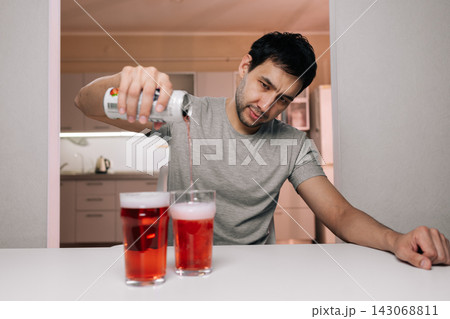 Bearded young man pouring alcohol into glasses at home, isolated and distressed, depicting loneliness, dependence and emotional struggle of substance abuse and self medication. Bearded young man pouring alcohol into glasses at home, isolated and distressed, depicting loneliness, dependence and emotional struggle of substance abuse and self medication. 143068811