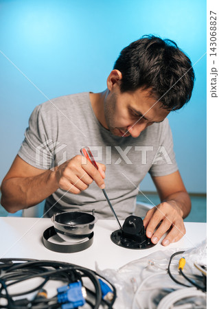 Vertical portrait of technician using screwdriver to assemble, repair dome security camera, working on components, wiring on white table against blue background, focusing on electronics maintenance. Vertical portrait of technician using screwdriver to assemble, repair dome security camera, working on components, wiring on white table against blue background, focusing on electronics maintenance. 143068827