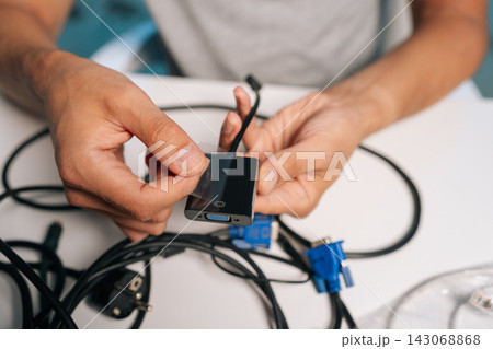 Close-up hands of unrecognizable person peeling transparent protective film from new black video adapter, preparing device for connection amidst tangle of cables and connectors on white desk. Close-up hands of unrecognizable person peeling transparent protective film from new black video adapter, preparing device for connection amidst tangle of cables and connectors on white desk. 143068868