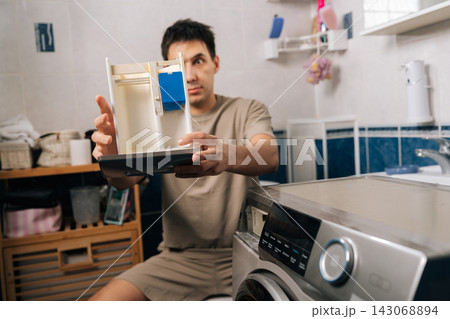 Puzzled man expressing disgust with broken washing machine dispenser, holding dirty powder tray showing repair issue and laundry appliance problem. Concept of household chores. Puzzled man expressing disgust with broken washing machine dispenser, holding dirty powder tray showing repair issue and laundry appliance problem. Concept of household chores. 143068894