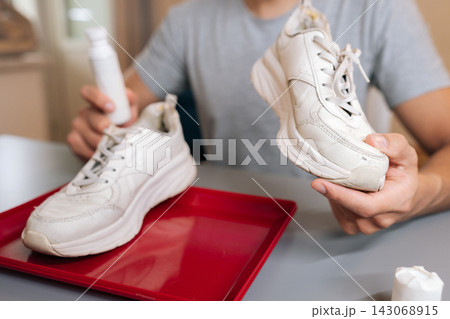 Close-up cropped shot of unrecognizable man holding shoe cleaner and white sneaker, preparing for cleaning and maintenance, focusing on domestic tasks and product use for footwear restoration. 143068915
