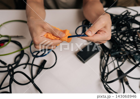 Close-up of person hands cleaning blue cable with an orange cloth, surrounded by a chaotic assortment of black and green audio, signal, and power cables on a white table. 143068946