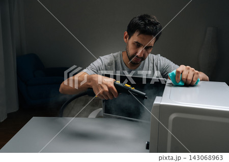 Male cleaning microwave oven using handheld steam cleaner, applying hot vapor to disinfect and remove grime, maintaining hygiene in kitchen appliance with modern tools. Concept of kitchen hygiene. 143068963