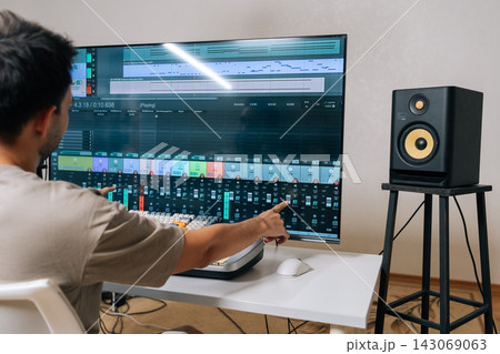 Back view of sound engineer male creating new music in home studio, arranging tracks and mixing audio levels with midi keyboard and software on a large display, showcasing modern music production. 143069063
