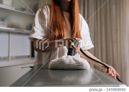 Close-up view of housewife cleaning kitchen microwave or appliance top with steam cleaner and white microfiber pad, producing visible steam to sanitize and deep clean surface. 143069093