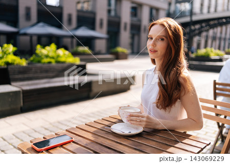 Pretty redhead woman sitting at wooden table in outdoor cafe, holding coffee cup and looking at camera, embodying relaxed and happy urban lifestyle under sun. Pretty redhead woman sitting at wooden table in outdoor cafe, holding coffee cup and looking at camera, embodying relaxed and happy urban lifestyle under sun. 143069229
