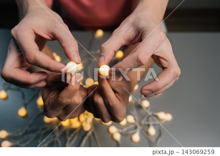 Person hands arranging decorative luminous garland lights providing cozy ambiance, preparing for celebration or home decoration project, close-up, cropped shot. 143069259