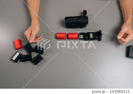 High-angle view of filmmaker loading rechargeable battery into black camera grip accessory, preparing for extended shooting sessions and enhanced vertical handling performance, close-up, cropped shot. High-angle view of filmmaker loading rechargeable battery into black camera grip accessory, preparing for extended shooting sessions and enhanced vertical handling performance, close-up, cropped shot. 143069315