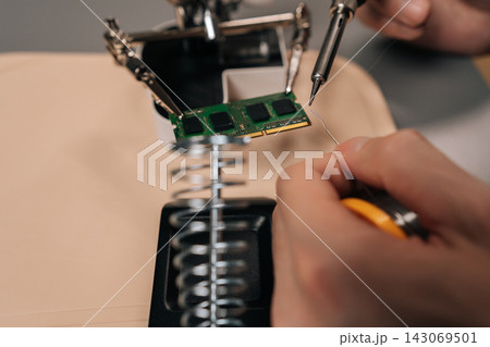 Male hands meticulously soldering an electronic component on a ram module, utilizing a soldering iron and helping hands tool for precise electronics repair and assembly. 143069501