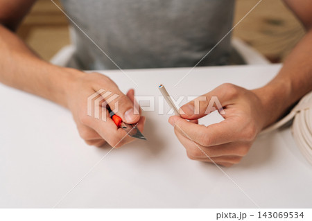 Top view of electrician hands splitting communication cable with utility knife sitting at white table, preparing data wires for connection and installation, performing electrical repair work. Top view of electrician hands splitting communication cable with utility knife sitting at white table, preparing data wires for connection and installation, performing electrical repair work. 143069534