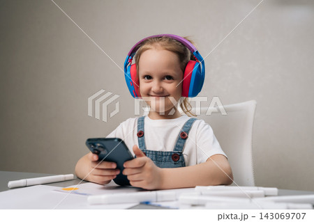 Studio portrait of young girl smiling while wearing colorful headphones and holding smartphone, concept of childhood, leisure, modern technology, online content consumption in bright setting. Studio portrait of young girl smiling while wearing colorful headphones and holding smartphone, concept of childhood, leisure, modern technology, online content consumption in bright setting. 143069707