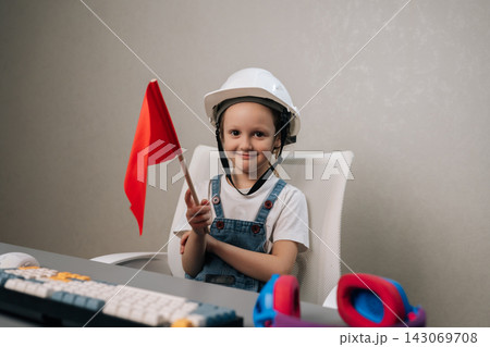 Young child wearing white safety helmet and blue overalls, smiling looking at camera while holding red warning flag at desk with keyboard, symbolizing safety and future careers. 143069708