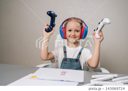 Studio portrait of young girl with big headphones smiling, lifting two game controllers in air, expressing joy and excitement about video gaming and childhood entertainment. Studio portrait of young girl with big headphones smiling, lifting two game controllers in air, expressing joy and excitement about video gaming and childhood entertainment. 143069724