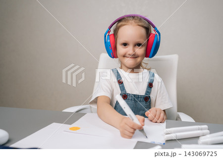 Smiling young child wearing colorful headphones, sitting at desk and drawing on paper with marker, actively engaged in creative and educational activity. Smiling young child wearing colorful headphones, sitting at desk and drawing on paper with marker, actively engaged in creative and educational activity. 143069725