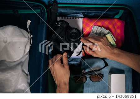 Close-up top view of traveler person packing clothes, camera, cash, sunglasses and a power bank into a suitcase, hands arranging travel essentials in preparation for a vacation or weekend trip. Close-up top view of traveler person packing clothes, camera, cash, sunglasses and a power bank into a suitcase, hands arranging travel essentials in preparation for a vacation or weekend trip. 143069851