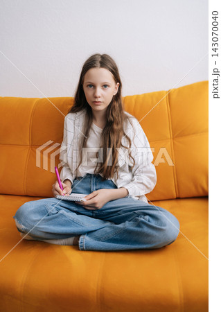 Vertical portrait of young female student sitting cross legged on comfortable bright yellow couch, holding pen and notebook, focusing on studies and completing homework in casual home environment. Vertical portrait of young female student sitting cross legged on comfortable bright yellow couch, holding pen and notebook, focusing on studies and completing homework in casual home environment. 143070040