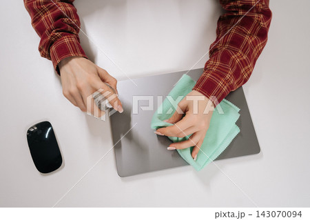 Top down view of woman hands cleaning silver laptop with sanitizer spray and green cloth. Concept of hygiene, tech care, and workplace sanitation. Clean, minimalist composition on white desk. Top down view of woman hands cleaning silver laptop with sanitizer spray and green cloth. Concept of hygiene, tech care, and workplace sanitation. Clean, minimalist composition on white desk. 143070094