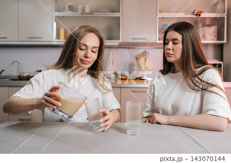 Two young women in kitchen. One pours homemade fruit smoothie from blender into glasses. Friend watches with skeptical expression. Concept for healthy lifestyle, diet, friendship, morning routine. 143070144