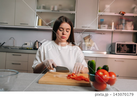 Concentrated young woman with eyes closed slices red tomatoes on cutting board. She wears gloves while preparing healthy food in her home kitchen. Domestic lifestyle and vegetarian meal prep concept. Concentrated young woman with eyes closed slices red tomatoes on cutting board. She wears gloves while preparing healthy food in her home kitchen. Domestic lifestyle and vegetarian meal prep concept. 143070179