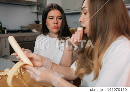 Two young women in kitchen. One slices banana on cutting board while looking with judgmental expression at her friend eating piece of fruit. Candid lifestyle scene showing friendship and diet. 143070180