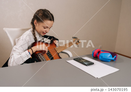 Young girl with pigtails focused on playing bass guitar, following online lesson on smartphone, developing new musical skills and a creative hobby at home. 143070195