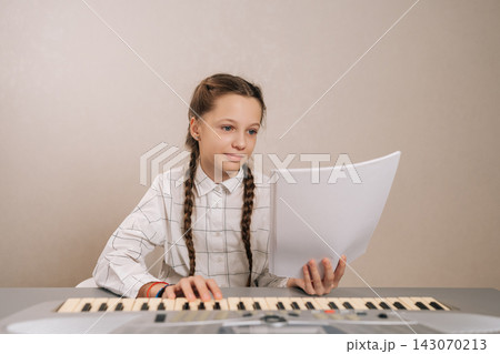 Portrait of talented adorable child girl with pigtails engaging in music lesson, concentrating on score presented on blank sheet of paper while pressing keys on synthesizer, acquiring new abilities. Portrait of talented adorable child girl with pigtails engaging in music lesson, concentrating on score presented on blank sheet of paper while pressing keys on synthesizer, acquiring new abilities. 143070213