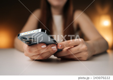 Female hands holding modern smartphone, browsing internet or using social media apps, enjoying mobile technology sitting at white table in dark comfortable setting, close-up, bokeh light. Female hands holding modern smartphone, browsing internet or using social media apps, enjoying mobile technology sitting at white table in dark comfortable setting, close-up, bokeh light. 143070222