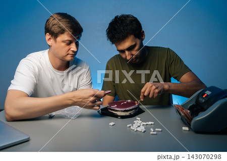 Portrait of two young men sitting at table eating cake laced with drugs, pills scattered on table near money counter, suggesting illegal activity and celebration of ill-gotten gains. Portrait of two young men sitting at table eating cake laced with drugs, pills scattered on table near money counter, suggesting illegal activity and celebration of ill-gotten gains. 143070298
