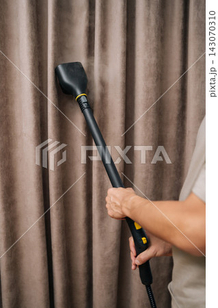 Vertical cropped shot of person using steam cleaner wand on large fabric curtains, performing professional textile cleaning and sanitizing for home hygiene, neatness, fresh drapes. Vertical cropped shot of person using steam cleaner wand on large fabric curtains, performing professional textile cleaning and sanitizing for home hygiene, neatness, fresh drapes. 143070310