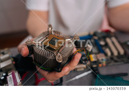 Hand holding CPU mounted on heatsink above computer motherboard, ready for installation or repair close-up of technician performing hardware maintenance and upgrade. Hand holding CPU mounted on heatsink above computer motherboard, ready for installation or repair close-up of technician performing hardware maintenance and upgrade. 143070359
