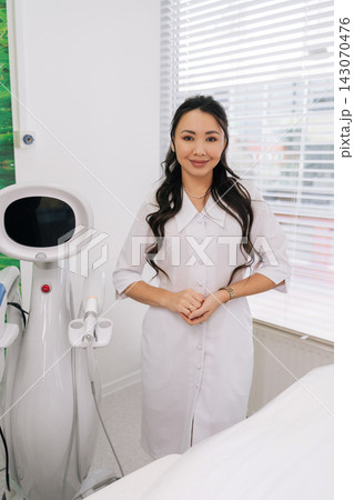 Positive Asian cosmetologist in white uniform standing next to advanced SMAS lifting machine in clean beauty clinic, preparing for procedure offering skincare and anti-aging treatments. Positive Asian cosmetologist in white uniform standing next to advanced SMAS lifting machine in clean beauty clinic, preparing for procedure offering skincare and anti-aging treatments. 143070476