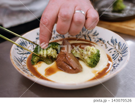 Serving food. Portioned dish. Chef male hand adding with tweezers grilled green broccoli to sauce polenta. Hearty tasty restaurant recipe. 143070526