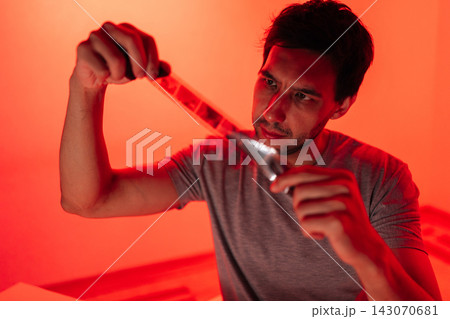 Young man examining photographic negative film strip with flashlight, focusing on details in red lit darkroom, connecting with traditional analog photography. Young man examining photographic negative film strip with flashlight, focusing on details in red lit darkroom, connecting with traditional analog photography. 143070681