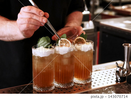 Decorating cocktail. Alcoholic beverage. Male barkeeper hand adding dried orange slice in aromatic liquid with frothy top mint leaves. Decorating cocktail. Alcoholic beverage. Male barkeeper hand adding dried orange slice in aromatic liquid with frothy top mint leaves. 143070714