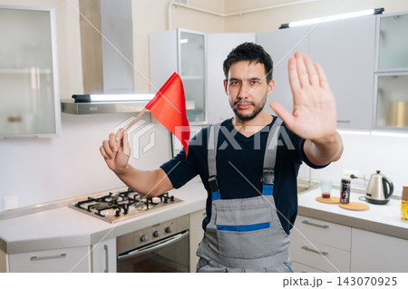 Repairman in uniform holding red flag and gesturing stop with hand in modern kitchen, signaling caution or danger during appliance or gas hob maintenance.. Repairman in uniform holding red flag and gesturing stop with hand in modern kitchen, signaling caution or danger during appliance or gas hob maintenance.. 143070925