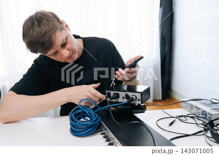 Focused man sets up audio equipment, connecting cables to sound interface with smartphone. Bright, natural lighting in modern home studio. Technical concept for music production or sound engineering. Focused man sets up audio equipment, connecting cables to sound interface with smartphone. Bright, natural lighting in modern home studio. Technical concept for music production or sound engineering. 143071005