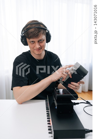 Young male music producer wearing headphones smiles while adjusting electronic synthesizer at desk. Bright, high key lighting in modern home recording studio. Creative hobby or profession. Young male music producer wearing headphones smiles while adjusting electronic synthesizer at desk. Bright, high key lighting in modern home recording studio. Creative hobby or profession. 143071016
