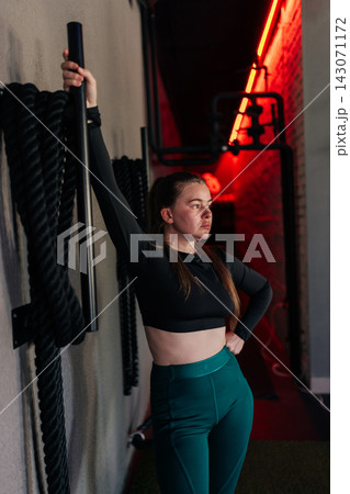 Vertical portrait of sporty woman in sportswear feeling determined and strong, preparing for challenging workout session leaning on equipment in gym with red accent lighting. Vertical portrait of sporty woman in sportswear feeling determined and strong, preparing for challenging workout session leaning on equipment in gym with red accent lighting. 143071172