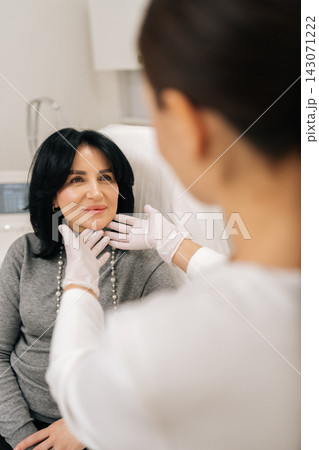 Close-up back view of professional female cosmetologist wearing gloves talking to mature woman patient in chair and examining facial skin before acosmetic procedure in modern aesthetic clinic. 143071222