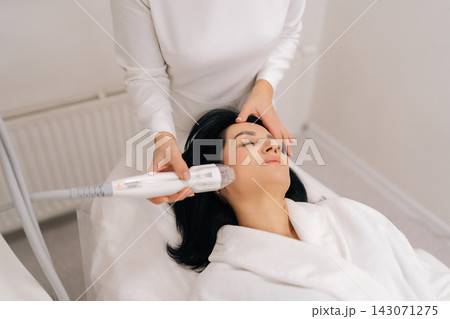 Top view of relaxed adult woman receiving innovative rf microneedling facelift procedure from skilled esthetician, modern hardware cosmetology method for skin tightening and wrinkle reduction. 143071275