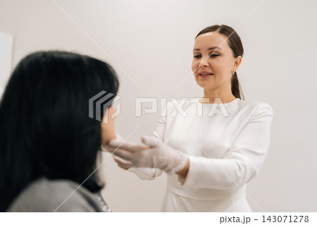 Portrait of professional female cosmetologist wearing gloves talking to mature woman patient in chair and examining facial skin before acosmetic procedure in modern aesthetic clinic. 143071278