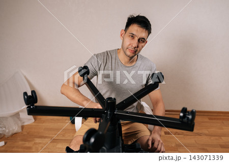 Portrait of young man assembling ergonomic office chair from flat-pack kit at home workspace, attaching wheels and components in focused diy setup for remote work, looking at camera. Portrait of young man assembling ergonomic office chair from flat-pack kit at home workspace, attaching wheels and components in focused diy setup for remote work, looking at camera. 143071339