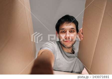 Curious man opens cardboard box looking inside. Unique low angle perspective from within package. Concept for delivery, ecommerce, moving, receiving gift. Neutral background focuses on emotion. Curious man opens cardboard box looking inside. Unique low angle perspective from within package. Concept for delivery, ecommerce, moving, receiving gift. Neutral background focuses on emotion. 143071691