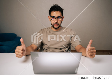 Portrait of serious bearded businessman wearing glasses, sitting at desk, working remotely from home showing thumbs up gesture of approval, showcasing success and satisfaction in career. Portrait of serious bearded businessman wearing glasses, sitting at desk, working remotely from home showing thumbs up gesture of approval, showcasing success and satisfaction in career. 143071720