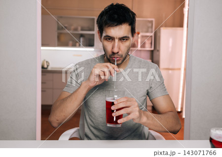 Young man sitting at white table drinking red beverage with straw, looking directly at camera with focused expression. Calm, everyday lifestyle scene in modern home interior. Refreshment concept. Young man sitting at white table drinking red beverage with straw, looking directly at camera with focused expression. Calm, everyday lifestyle scene in modern home interior. Refreshment concept. 143071766