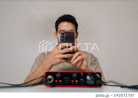 Front view of sound engineer using smartphone with headphones and external audio interface at home desk, focused on digital music production, streaming or podcasting in relaxed indoor setting. 143071797