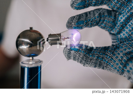 Closeup shot of scientist hand in protective glove holding glowing light bulb near high frequency coil, demonstrating wireless electricity transmission and energy innovation in physics experiment. Closeup shot of scientist hand in protective glove holding glowing light bulb near high frequency coil, demonstrating wireless electricity transmission and energy innovation in physics experiment. 143071908