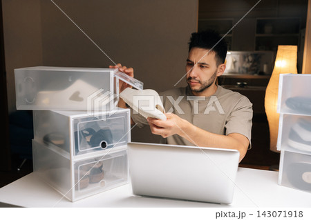 Focused male entrepreneur organizing shoe inventory at home, managing online store and e-commerce business using laptop, ensuring efficient storage, preparing for shipping. Concept of small business. Focused male entrepreneur organizing shoe inventory at home, managing online store and e-commerce business using laptop, ensuring efficient storage, preparing for shipping. Concept of small business. 143071918